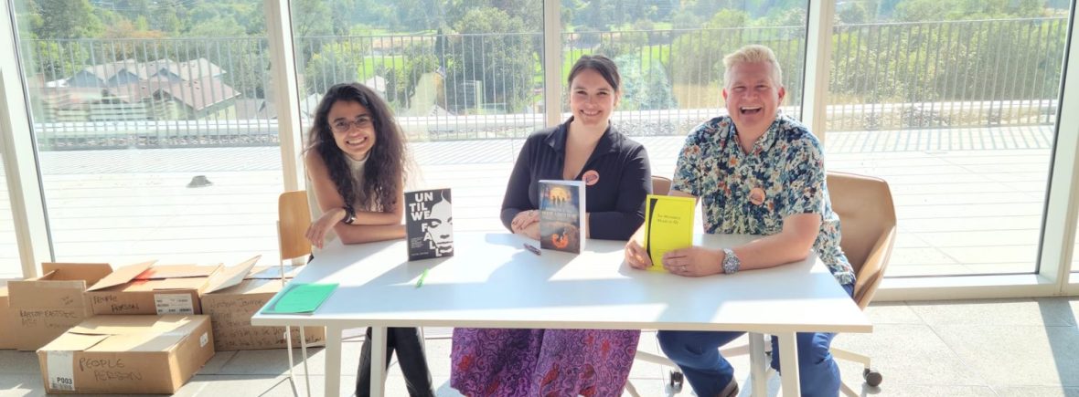 Three authors pose at a white table with their books. A wide window in the background shows the view of mountains and trees.